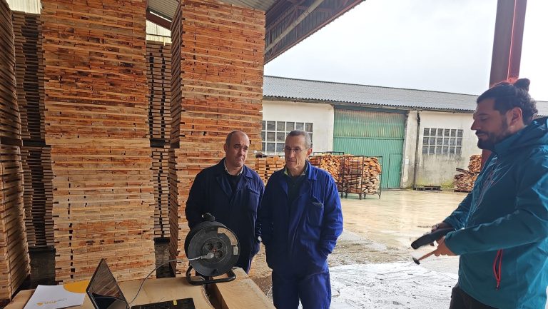 Tres personas en un aserradero navarro observan un equipo técnico sobre una mesa con ordenadores y sensores, rodeados de pilas de madera de haya.