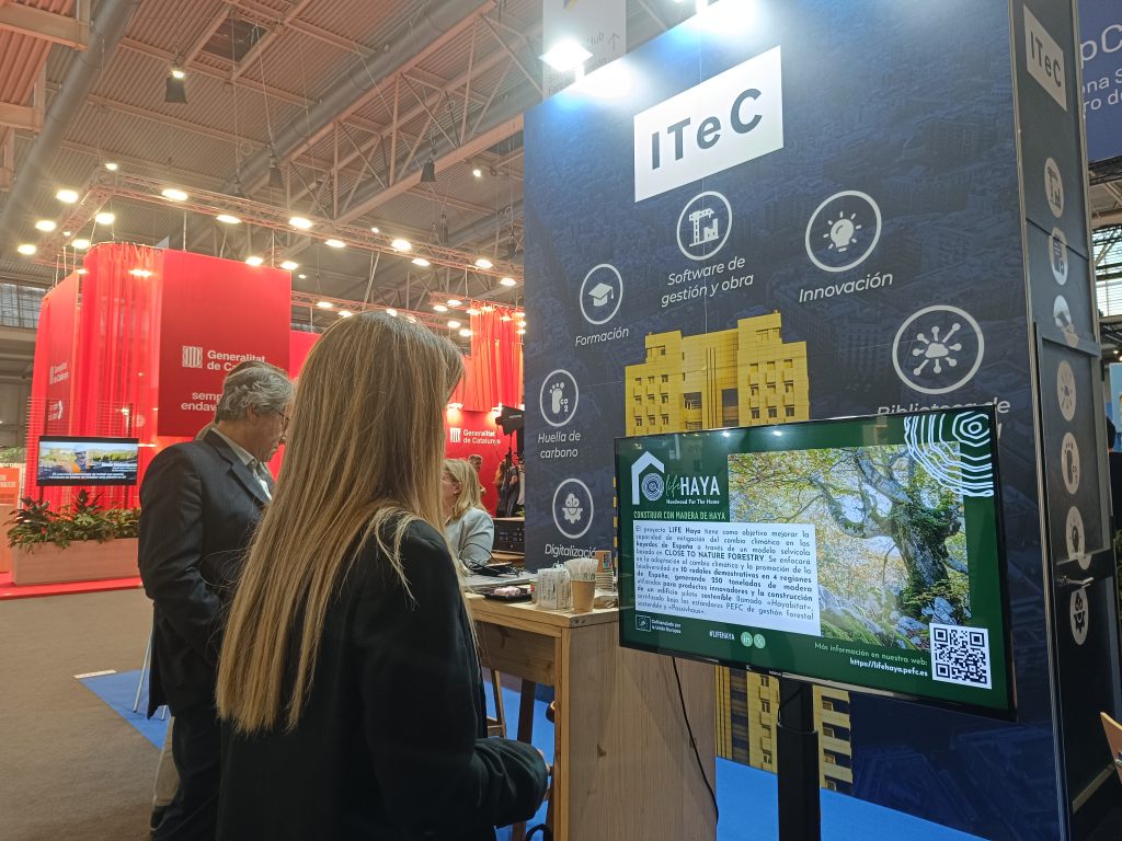 Dos personas, un hombre y una mujer, observan una pantalla en el stand de ITeC en Construmat 2024. La pantalla muestra información sobre el proyecto LIFE HAYA, incluyendo un gran árbol y texto.
