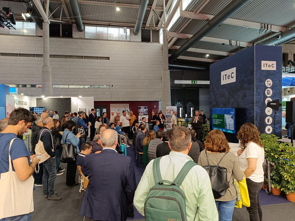 Asistentes escuchando una presentación en el stand del ITeC durante la feria Construmat 2024, con mención al proyecto LIFE HAYA en pantalla.