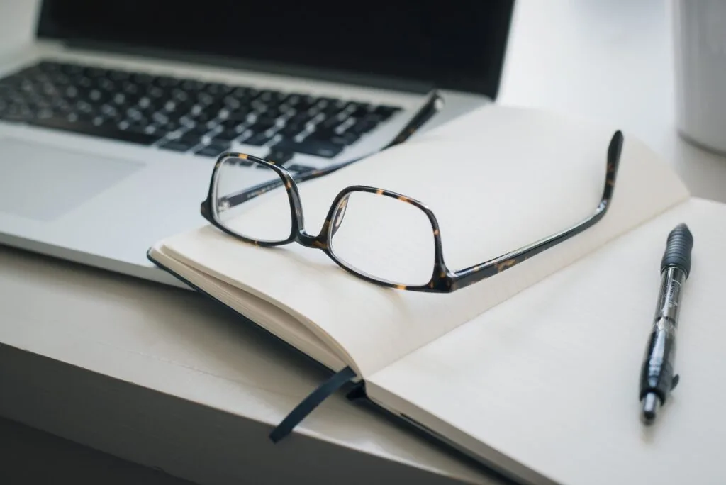 Cuaderno abierto con gafas y boligrafo junto a un portatil encendido